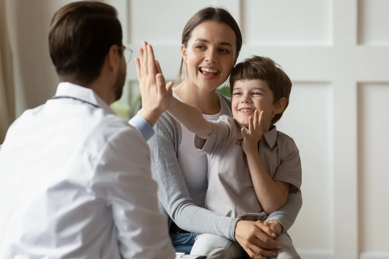 Glücklicher süßer kleiner Junge, der dem Kinderarzt High Five gibt, freundlicher Arzt, der Vorschulpatienten bei Arzttermin begrüßt, gutes Untersuchungsergebnis, junge Mutter und Sohn besuchen Hausarzt im Krankenhaus